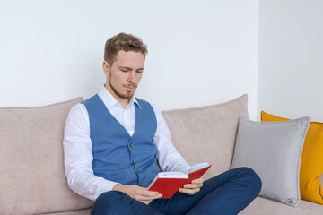 Focused young business man sits on couch with notebook in his hand and reads and analyzes an action plan for development at work in a blue suit. Caucasian businessman in the office on the couch