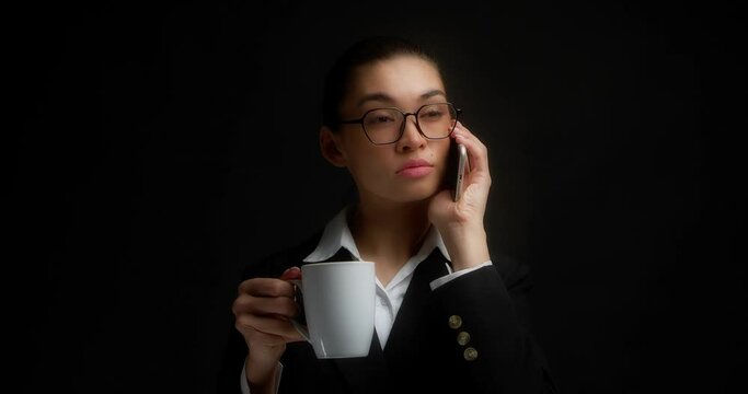 Business Woman With Glasses And Long Brown Hair Is Standing On A Black Background In Office Clothes, Talking Seriously On A Mobile Phone And Holding A White Coffee Mug In Her Hands.