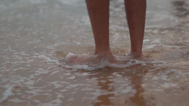 Low angle shot of legs of an Indian man standing in the water of the sea waves of the beach at Baga beach in Goa, India. Man soaks his legs on the beach water. Man enjoying his summer holidays. 