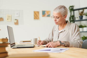 Concentrated middle-aged businesswoman with short hair sitting at desk in cozy office and working...