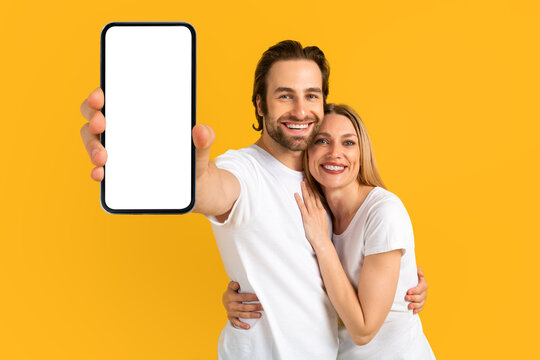 Happy young caucasian male and female in white t-shirts hugging, show smartphone with blank screen