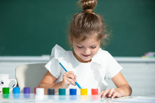 Little funny artist painting, drawing art. Child girl draws in classroom sitting at a table, having fun on school blackboard background.