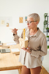 Stylish middle-aged businesswoman in eyeglasses eating noodles from box while having lunch break in office