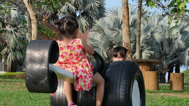 Cute kids playing on the outdoor playground. Little sisters sit on a see saw made of old tires in the park. Healthy summer activity for children.