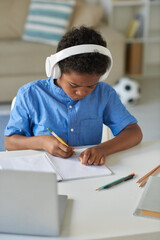 Concentrated black boy in wireless headphones sitting at table in living room and performing school...