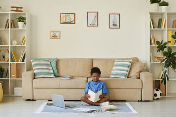 Fototapeta premium Concentrated black boy witting with crossed legs on floor and reading textbook while preparing for school lesson in living room