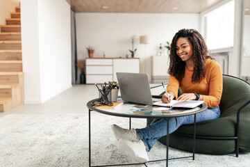 Smiling woman sitting at table, using computer writing in notebook