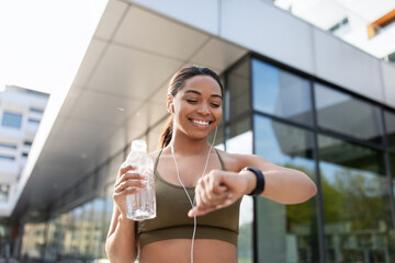 Black woman looking at smartwatch, tracking fitness activity, listening to music, drinking water on...
