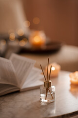 Liquid home fragrance in diffuser with open paper book on coffee table in bedroom indoors close up over scented candle. Cozy and hygge atmosphere. Aromatherapy.