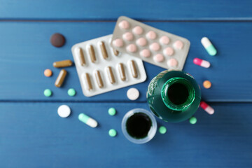 Bottle of cough syrup, measuring cup and pills on blue wooden background, flat lay