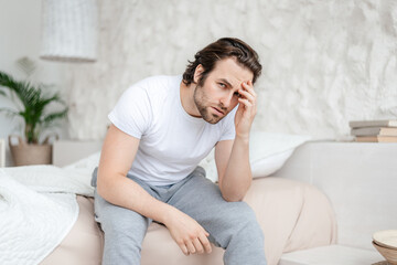 Sad millennial caucasian man with stubble sits on white bed, presses his hands to head and suffers from migraine