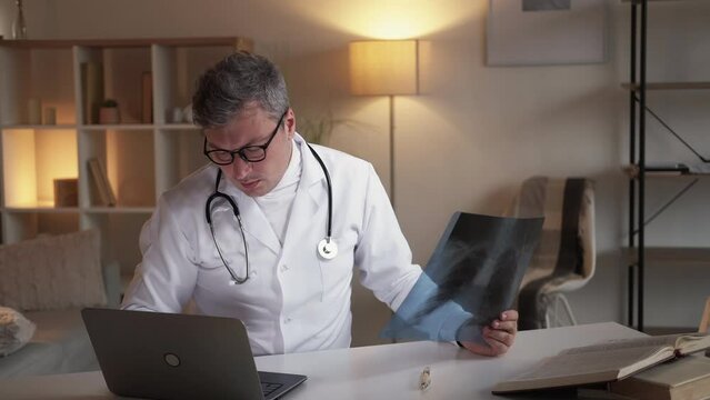 Patient diagnosis. Male doctor. Computer report. Considering man holding x-ray photo of lungs typing data on laptop sitting desk light room interior.