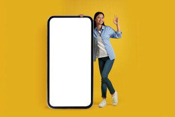 Satisfied young chinese woman in casual show ok sign with hand, stands near huge phone with empty screen - obrazy, fototapety, plakaty