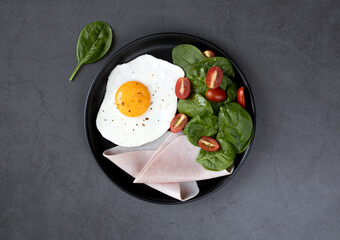Breakfast with scrambled eggs, spinach, ham and tomatoes in a black plate close-up on a dark background, top view