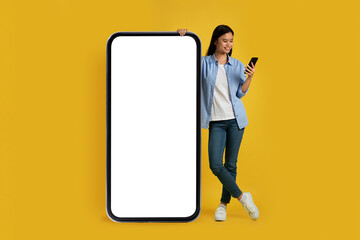 Happy young japanese female in casual look at smartphone, stands near huge phone with blank screen