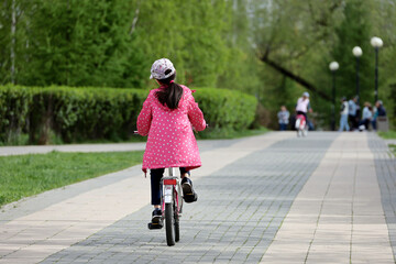 Kid girl riding on a bicycle on a path in a green park. Child cyclist, spring or summer leisure