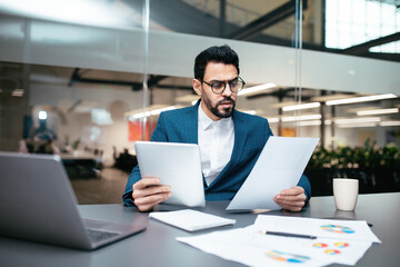 Serious busy handsome young muslim guy with beard in glasses, suit sits at table with computer and...
