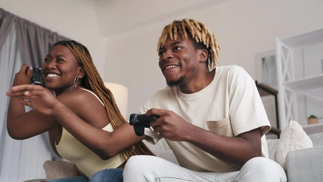 Home video game. Friends leisure. Fun weekend. Virtual challenge. Excited guy bothering girl trying to win playing with gamepad on couch in modern living room interior.