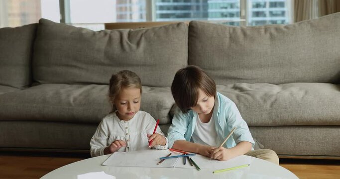 Cute focused siblings drawing with pencils at home. 5s girl her brother sit at table in modern living room engaged in painting, spend leisure enjoy favourite hobby. Kids growth and development concept