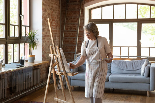 Focused older woman artist standing in loft workshop holds palette and paintbrush, drawing picture on canvas, feeling inspiration enjoy process. Creative hobby on retirement, activity for soul concept - Powered by Adobe