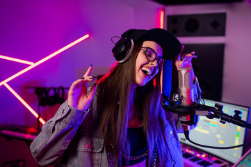 Beautiful stylish girl singing into a microphone in a recording studio with neon lights in a cap...