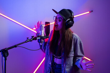 Beautiful stylish girl singing into a microphone in a recording studio with neon lights in a cap...