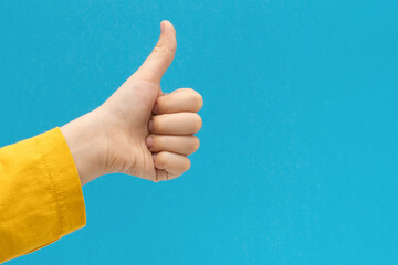 Close up child hand showing thumb up sign against blue background.