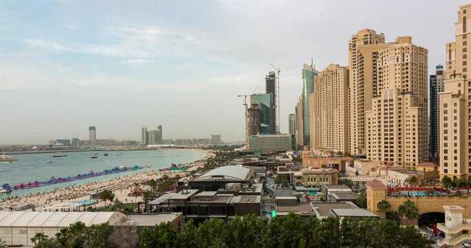 Aerial view of beach and tourists walking in JBR with skyscrapers on background day to night transition timelapse in Dubai, UAE. Waterfront with many activities and attractions, shops and restaurants