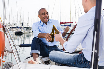 Two men in blue shirts and jeans with bare legs drinking beer and having fun on a yacht in the port