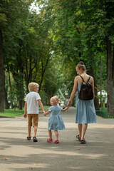 Young mother walks through summer park with her two children. Family walks along the alley in park. Back view. Vertical frame.