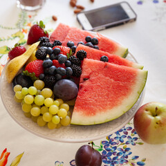 Sliced fruits and berries on a beautiful white plate with a phone on the back background. Watermelon, strawberry, blueberries, blackberry and grapes. Dietary fruit salad for breakfast