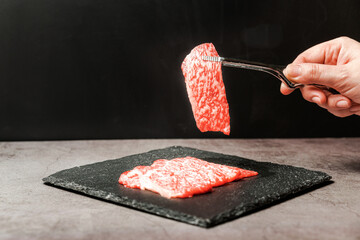 Premium Rare Slices sirloin Wagyu A5 beef with high-marbling texture pick up by bbq tongs with hand from stone plate. Served for Yakiniku, Sukiyaki and Shabu. Image with copy space.