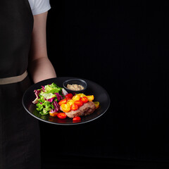 Grilled beef steak with grilled vegetables and salad leaves served on black plate on black background presentation in chef hands.
