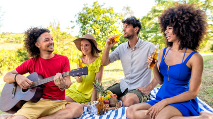 summer holidays outdoor picnic. multiracial group of friends having food and drinking beers laying on a blanket in a park garden. people happy hour enjoying together guitar music. lifestyle concept