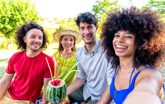 summer holidays outdoor picnic. multiracial group of friends having drink out of watermelon laying on a blanket in a park garden. people happy hour enjoying together making a selfie. lifestyle concept