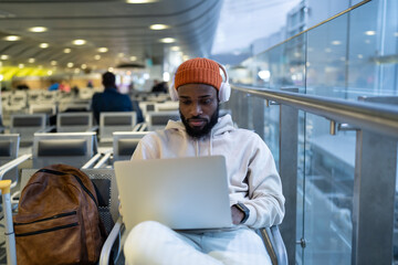 Young African American man sitting in hall of airport terminal using laptop, wear headphones and...