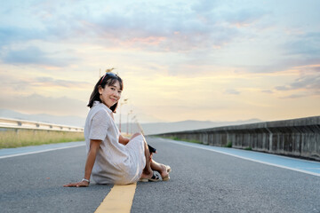 Woman traveler looking at the camera and sitting on the street of the dam with a sunset background on a vacation trip. A female Asian tourist enjoying nature on holiday. Freedom concept