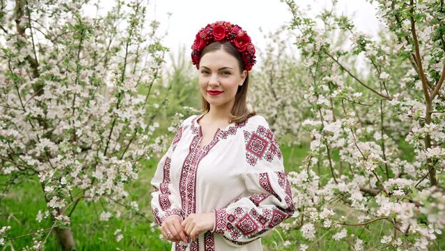 Portrait of pretty ukrainian woman in blossom garden. Girl in traditional embroidery vyshyvanka dress and red wreath. Ukraine freedom, springtime, national costume, victory in war.