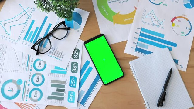 Top view of smartphone with green screen on the office desk. Application for business. Notebook, graphs, eyeglasses and plant on office table workspace. Chroma key.