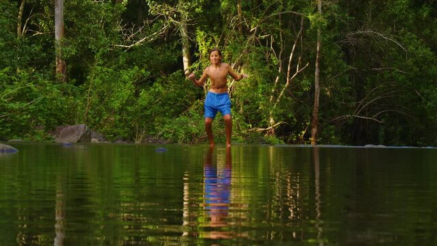 The boy dances cheerfully in the park against the backdrop of green trees. A beautiful reflection on the surface of the water of a dancing teenager. Water splash