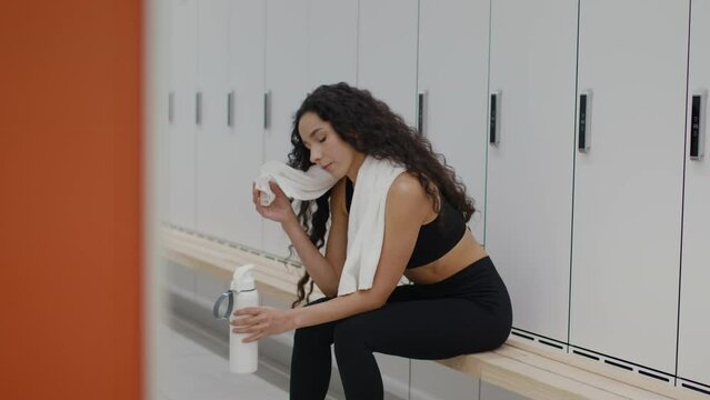 Young active sporty woman resting after hard workout at gym locker room, drinking water and wiping sweat from face