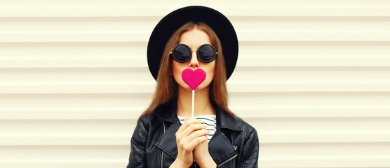 Portrait of stylish young woman with pink heart shaped lollipop posing wearing black round hat on...