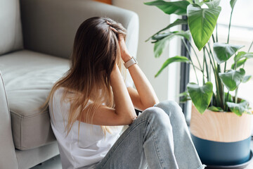 Unhappy young caucasian woman with blonde hair thinking about bad relationships problems, break up with boyfriend. Worried millennial girl sitting on floor in bedroom near chair and green plant alone - obrazy, fototapety, plakaty