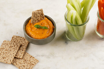 Paprika bell-pepper hummus dip looking like orange pumpkin puree, thin crackers with sesame seeds and carrot and cucumber cut in long strips and sticks on marble board