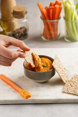 Female hand reaching for paprika bell-pepper hummus dip looking like orange pumpkin puree, thin crackers with sesame seeds and carrot and cucumber cut in long strips and sticks on marble board