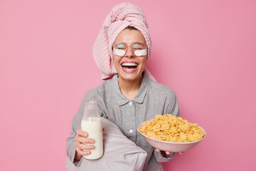 Positive young female model going to eat delcious healthy breakfast holds bowl of cornflakes and bottle of milk dressed in nightwear laughs happily has good mood isolated over pink background