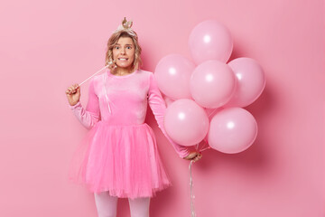 Obraz na płótnie Canvas Festive occasion and holiday concept. Beautiful European woman has worried facial expression wears crown and dress holds magic wand and bunch of inflated balloons isolated over pink background.