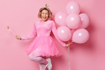 Obraz na płótnie Canvas Upbeat cheerful female princess wears festive dress dances carefree holds magic wand and bunch of inflated helium balloons has fun on party isolated over pink studio background. Monochrome shot