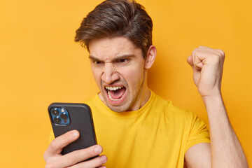 Outraged angry adult man screams loudly focused at smartphone screen clenches fist has argument...