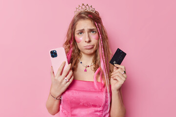 Unhappy long haired young woman holds mobile phone and credit card makes payment online wears dress...